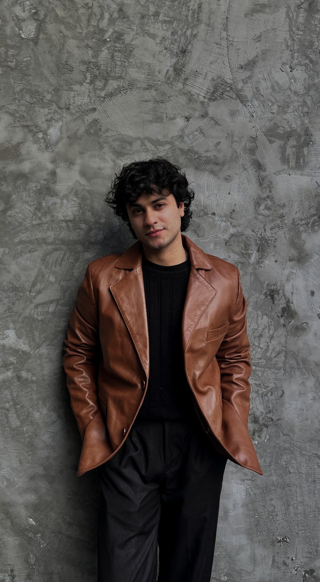 Man in a brown leather blazer, hands in pockets, against a concrete wall